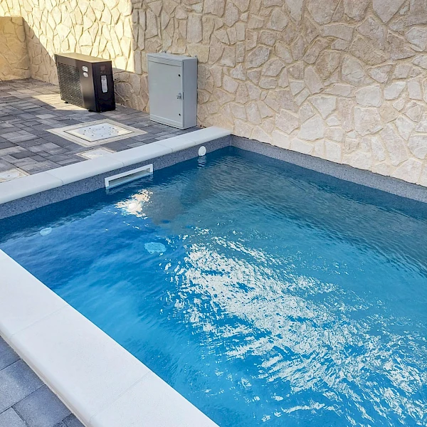 Napunjen vanjski bazen uz kameni zid, s modernom crnom inverter toplinskom pumpom za grijanje vode ugrađenom u pozadini