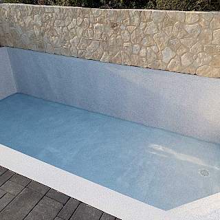 Prazan vanjski bazen sa svijetlo sivom armiranom folijom i vidljivim podnim slivnikom, smješten uz ukrasni kameni zid