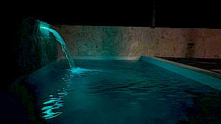 Intimni noćni kadar vanjskog bazena sa snažnim mlazom vode iz ugrađenog zidnog bazenskog slapa obasjanog suptilnom svjetlošću.