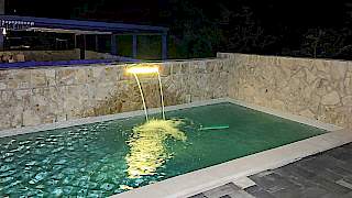 Noćni prikaz vanjskog bazena s ugrađenim osvijetljenim slapom u kamenom zidu koji stvara predivan vizualni vodeni efekt.