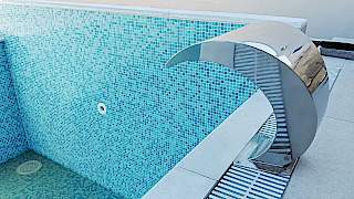 Elegantni sjajni inox bazenski slap postavljen na rub modernog vanjskog bazena s folijom u uzorku plavog mozaika.