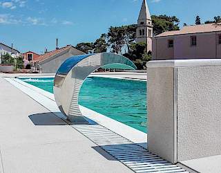 Sjajni inox bazenski slap postavljen uz rub vanjskog preljevnog bazena, s predivnim mediteranskim zvonikom u pozadini.