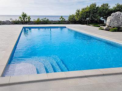 Moderni pravokutni vanjski bazen sa svijetlo plavom vodom, ugrađenim stepenicama i kamenim rubom uz more